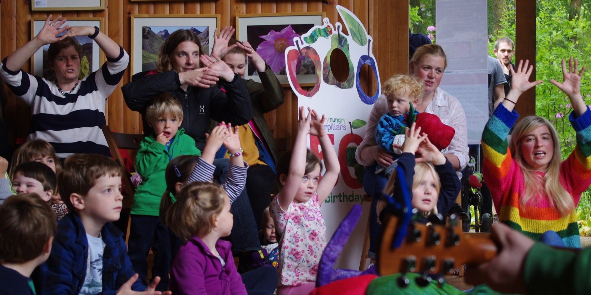 A photo of parents with children in a wooden room. They are all making hand signals with both hands and looking in the same direction.