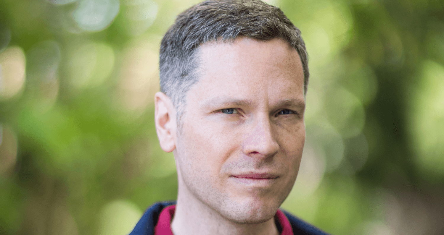 Allan Radcliffe. A man dressed in a red top with a blurred background of leaves.