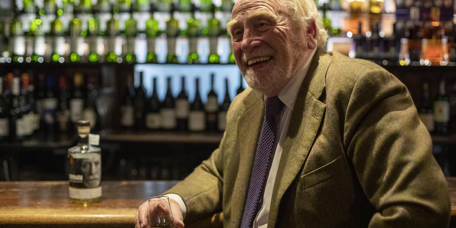 A man smiling sat at a bar with a dram of whisky in hand.