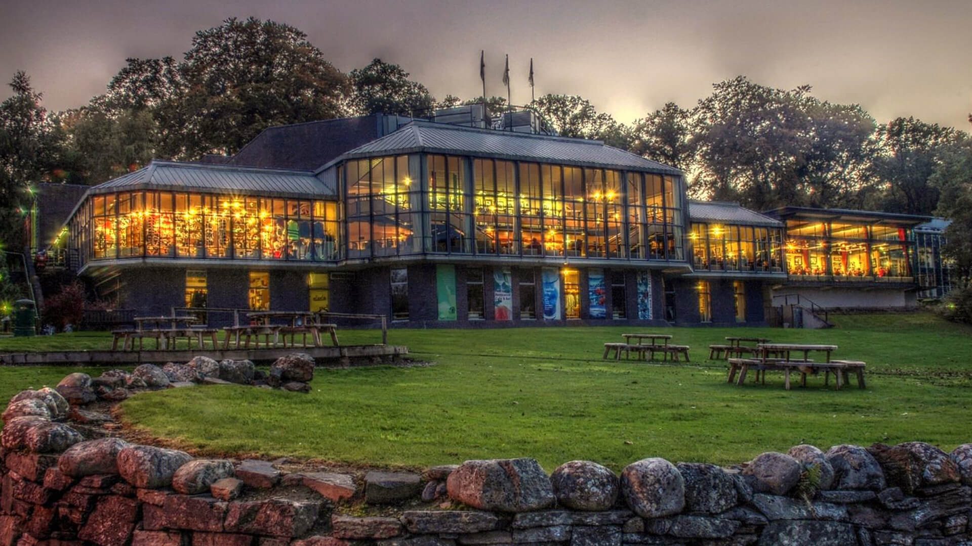 An image of the front of the main building of Pitlochry Festival Theatre with a full glass front and lights shining through each one, the ground outside is all covered in grass with wooden benches populating different parts and an evening sky above