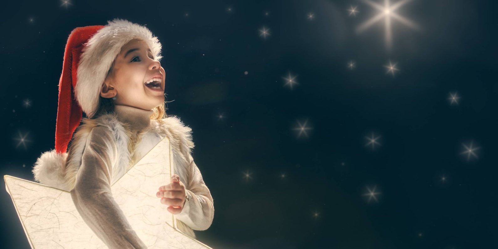 Girl in santa hat holding a glowing star looking up at star-filled sky