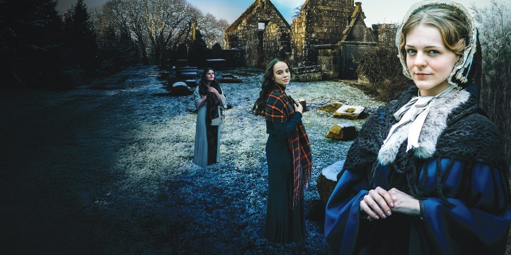 Three women in period dress standing in a frosty graveyard with church ruins in the background.