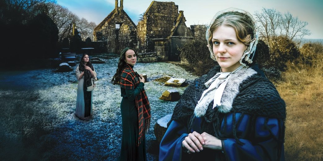 Three women in period dress standing in a frosty graveyard with church ruins in the background.