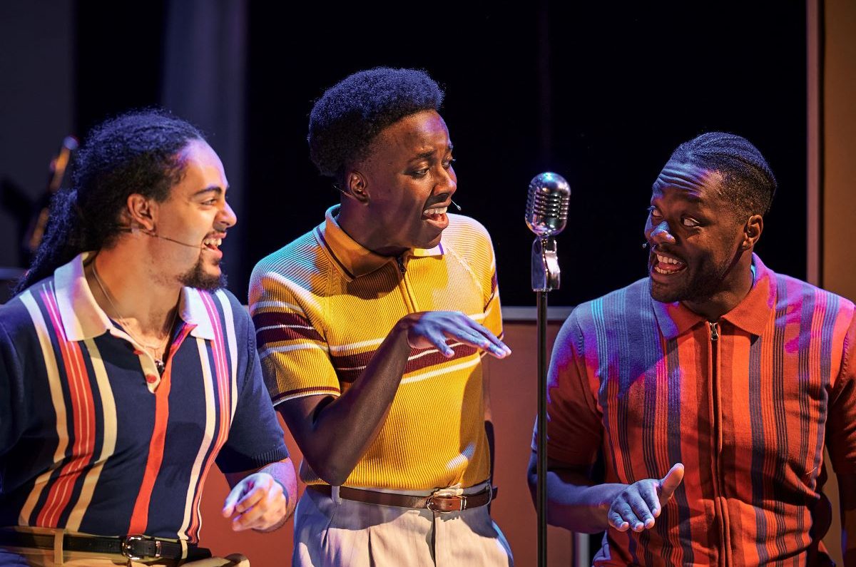 Three actors in costume singing round a chrome mic stand