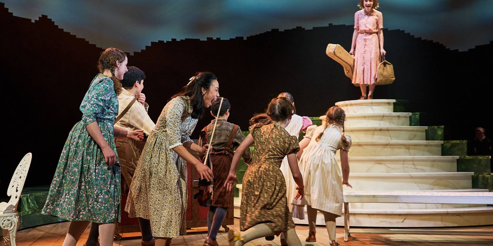 A play on stage with woman standing at top of the steps and a group of children in costume running towards her